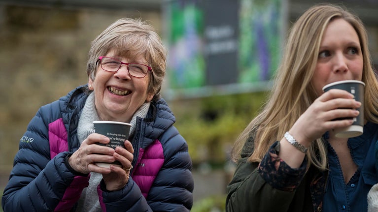 Two ladies in coats smile and laugh holding takeaway coffee cups